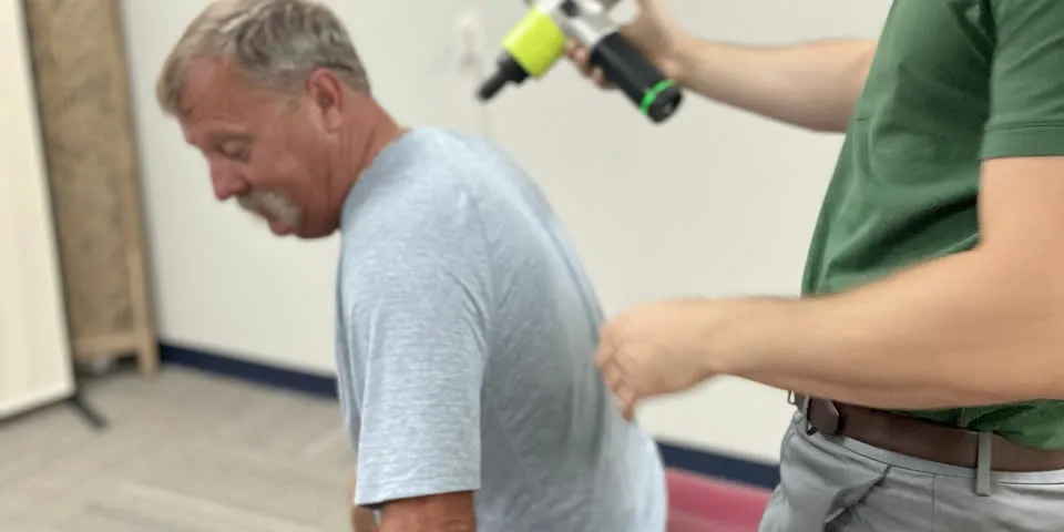 Man in a gray shirt standing beside a provider in a green polo who holds a handheld percussion massage device near his upper back