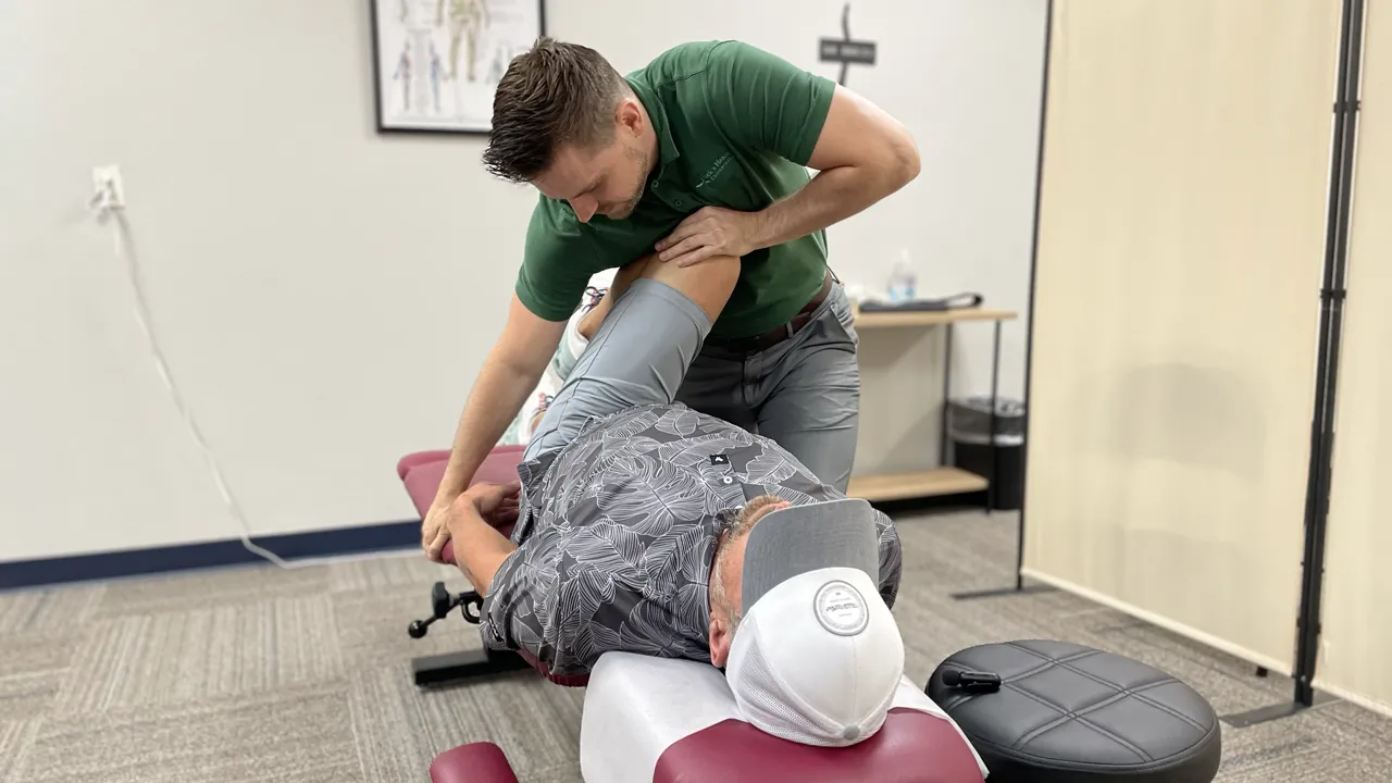 Provider in a green polo guiding a patient's bent leg across the body during a hip adjustment on a treatment table