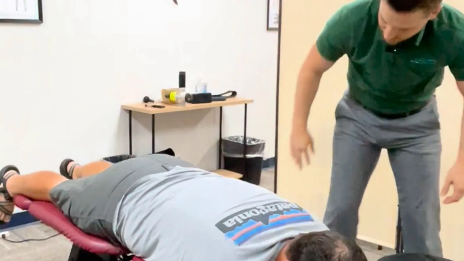 Person lying face down on a treatment table while a provider in a green polo stands beside the table