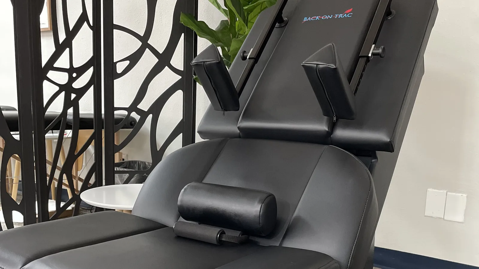 Empty Back-On-Trac spinal decompression chair in the clinic, with a decorative screen and a plant behind it